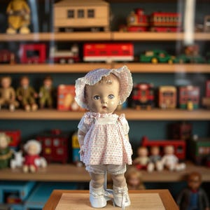 May include: A porcelain doll with a pink and white dress and a white bonnet. The doll has blue eyes and is standing on a wooden surface. The doll is surrounded by toy shelves with various toys.