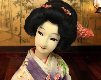 Puede incluir: Una muñeca japonesa geisha con una expresión serena. La muñeca tiene cabello negro peinado en un moño tradicional, adornado con un accesorio con estampado rojo y blanco. Viste un kimono colorido con estampados florales y geométricos.
