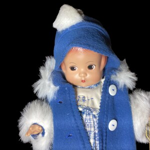 Op de afbeelding: Een porseleinen pop die een blauwe jas draagt met witte bontkraag en een witte hoed. De pop heeft een geschilderd gezicht met roze wangen en bruine ogen.
