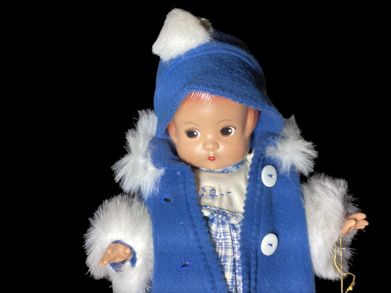 Op de afbeelding: Een porseleinen pop die een blauwe jas draagt met witte bontkraag en een witte hoed. De pop heeft een geschilderd gezicht met roze wangen en bruine ogen.