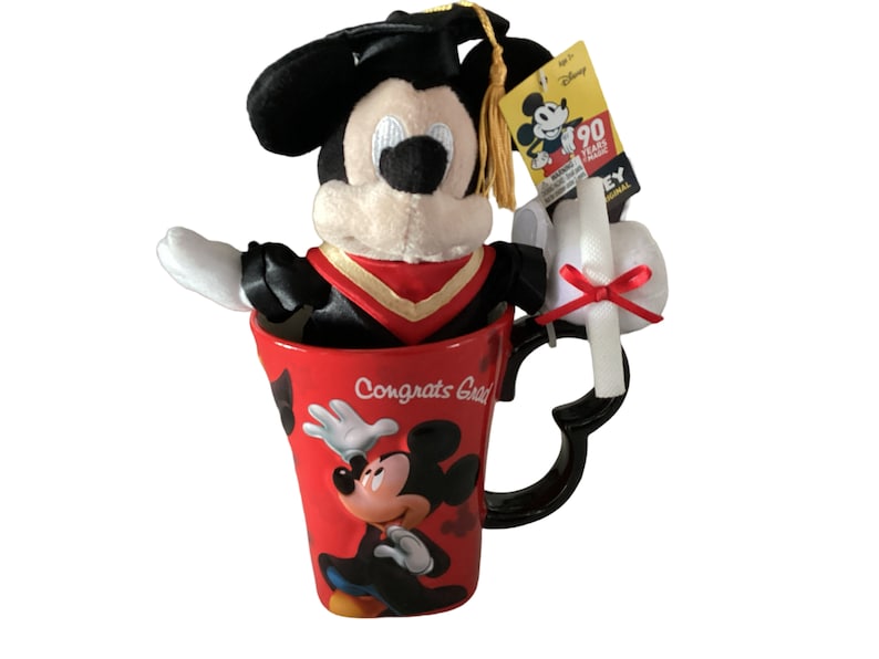 May include: A red ceramic mug with a black handle and a Mickey Mouse design. The mug says "Congrats Grad" and features a graduation cap. A plush Mickey Mouse wearing a graduation cap and gown is sitting inside the mug. A white graduation scroll with a red ribbon is attached to the mug.