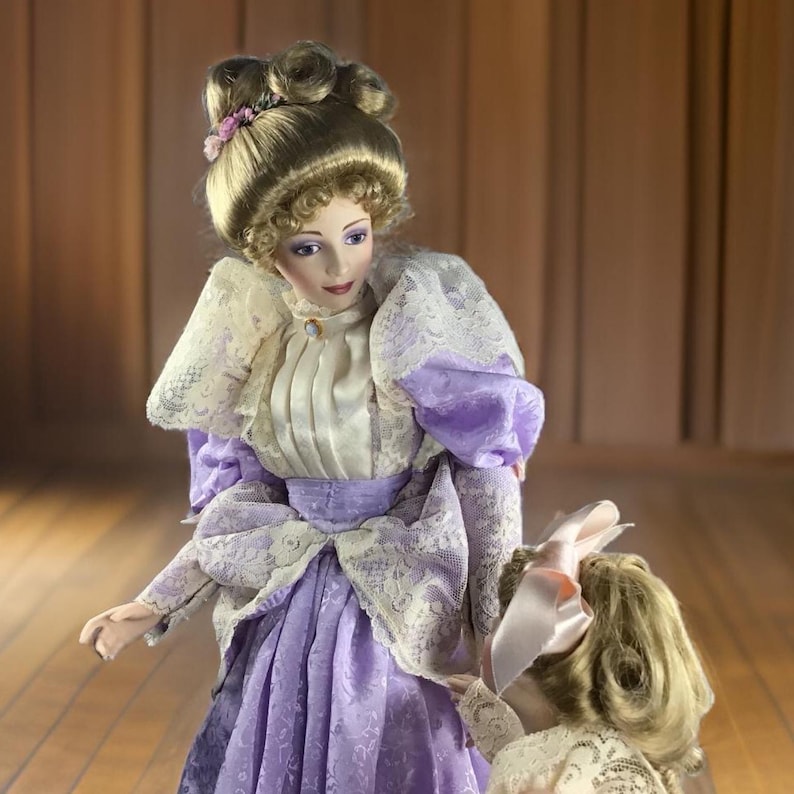 Puede incluir: Dos mu&ntilde;ecas de porcelana vestidas con ropa de inspiraci&oacute;n vintage. La mu&ntilde;eca m&aacute;s alta lleva un vestido lavanda con ribetes de encaje y una blusa blanca. La mu&ntilde;eca m&aacute;s baja lleva un vestido blanco con ribetes de encaje y un lazo rosa. Ambas mu&ntilde;ecas tienen el pelo rubio.