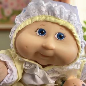Peut inclure: Une poupée Cabbage Patch Kid portant une robe jaune et blanche avec une bordure en dentelle blanche. La poupée a les yeux bleus et un teint rosé.
