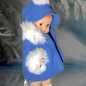 Op de afbeelding: Een porseleinen pop die een blauwe jas draagt met witte bontkraag en een witte pomponmuts. De pop draagt witte schaatsen en staat op een wit oppervlak.