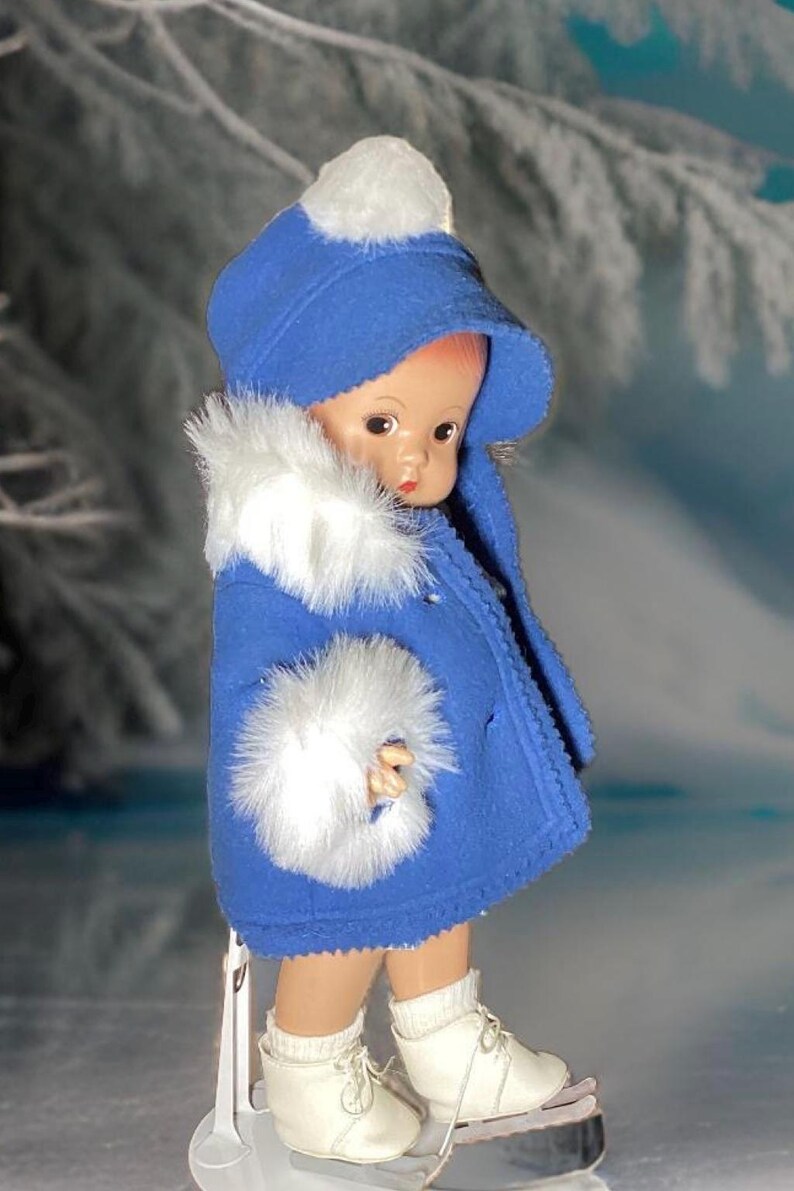 Op de afbeelding: Een porseleinen pop die een blauwe jas draagt met witte bontkraag en een witte pomponmuts. De pop draagt witte schaatsen en staat op een wit oppervlak.