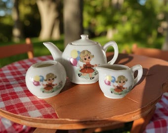 May include: A white ceramic tea set comprising a teapot, creamer jug, and sugar bowl. Each item is adorned with a cartoon girl holding balloons and wearing a red dress. The set is presented on a small wooden table.