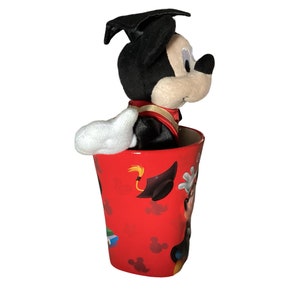 May include: A red ceramic mug with a graduation theme featuring Mickey Mouse wearing a graduation cap and gown. The mug has a black and white image of Mickey Mouse on the side.