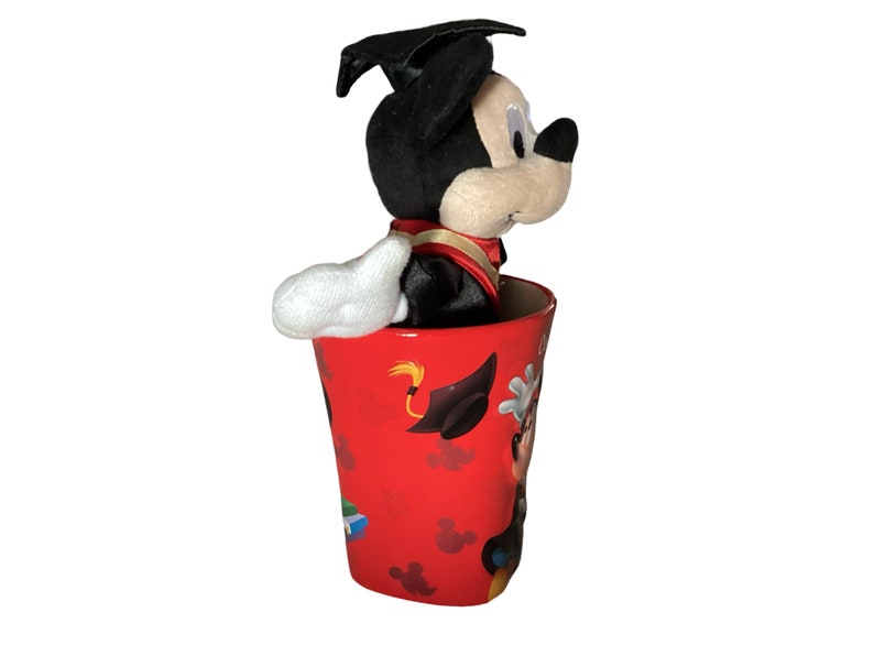 May include: A red ceramic mug with a graduation theme featuring Mickey Mouse wearing a graduation cap and gown. The mug has a black and white image of Mickey Mouse on the side.