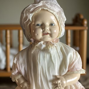 Puede incluir: Una muñeca bebé vintage con una expresión serena, que lleva un gorro blanco y un vestido rosa claro y blanco. La muñeca tiene ojos azules y mejillas rosadas. Una cuna de madera es visible en el fondo.