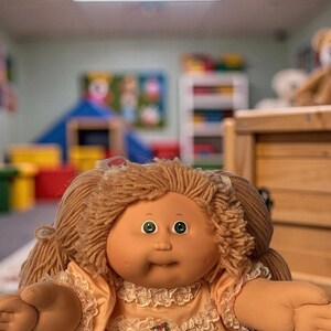 May include: A doll with light brown yarn hair styled in two pigtails, green eyes, and a small dimpled smile. The doll wears a peach-coloured dress featuring white lace trim around the neckline and bodice, with a small floral accent at the centre.