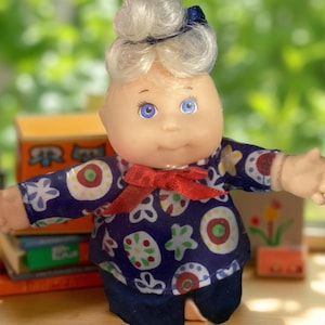Może przedstawiać: Lalka Cabbage Patch Kids z blond włosami i niebieskimi oczami, ubrana w niebiesko-białą koszulę w wzory z czerwoną kokardką i niebieskie spodnie.