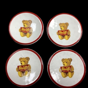 May include: Set of four white ceramic coasters with red trim. Each coaster features a brown teddy bear holding a red and yellow box with the text "Steiff" printed on it.