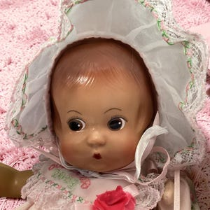 May include: A vintage baby doll with a painted face, wearing a pink dress and a white bonnet trimmed with lace and pink and green accents. The doll has large, dark eyes and a small red mouth. A pink rose adorns the dress.