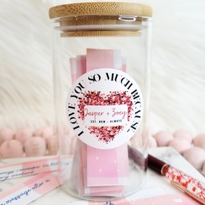 May include: A glass jar with a bamboo lid filled with pink paper slips. The jar has a white sticker with a pink heart and the text "I love you so much because Jasper + Zoey est. now. always".