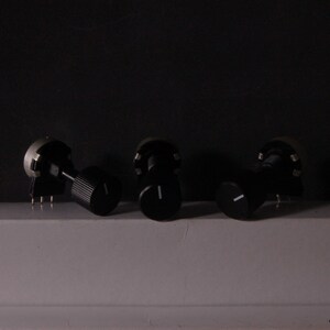 Peut inclure: Trois potentiom&egrave;tres noirs avec des lignes indicatrices blanches. Les boutons sont dispos&eacute;s sur une surface blanche sur fond sombre. Un bouton a une texture stri&eacute;e, les autres sont lisses.