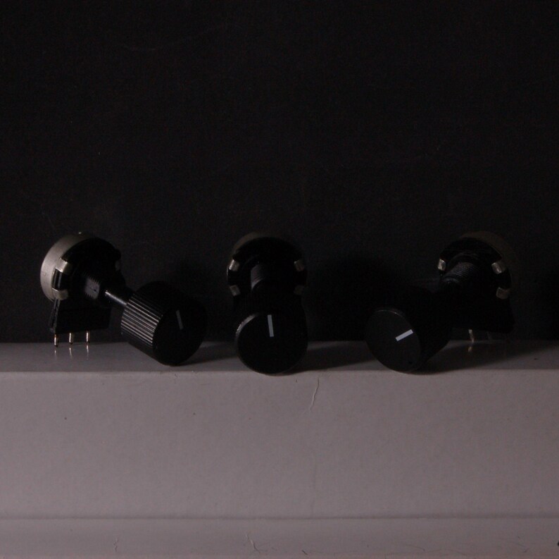 Peut inclure: Trois potentiom&egrave;tres noirs avec des lignes indicatrices blanches. Les boutons sont dispos&eacute;s sur une surface blanche sur fond sombre. Un bouton a une texture stri&eacute;e, les autres sont lisses.