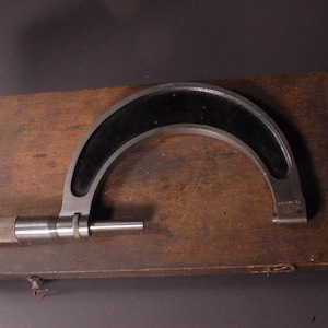 Könnte beinhalten: Ein Vintage-Mikrometer, ein Präzisionsmesswerkzeug, liegt auf einer Holzkiste. Das Messgerät hat einen schwarzen, gebogenen Rahmen und einen silbernen Messmechanismus. Die Holzkiste ist dunkelbraun.