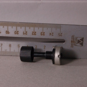 Peut inclure: Un bouton de potentiom&egrave;tre noir avec une surface stri&eacute;e, reli&eacute; &agrave; une tige noire et &agrave; une base argent&eacute;e. Une r&egrave;gle m&eacute;tallique avec des marquages en centim&egrave;tres est en arri&egrave;re-plan.