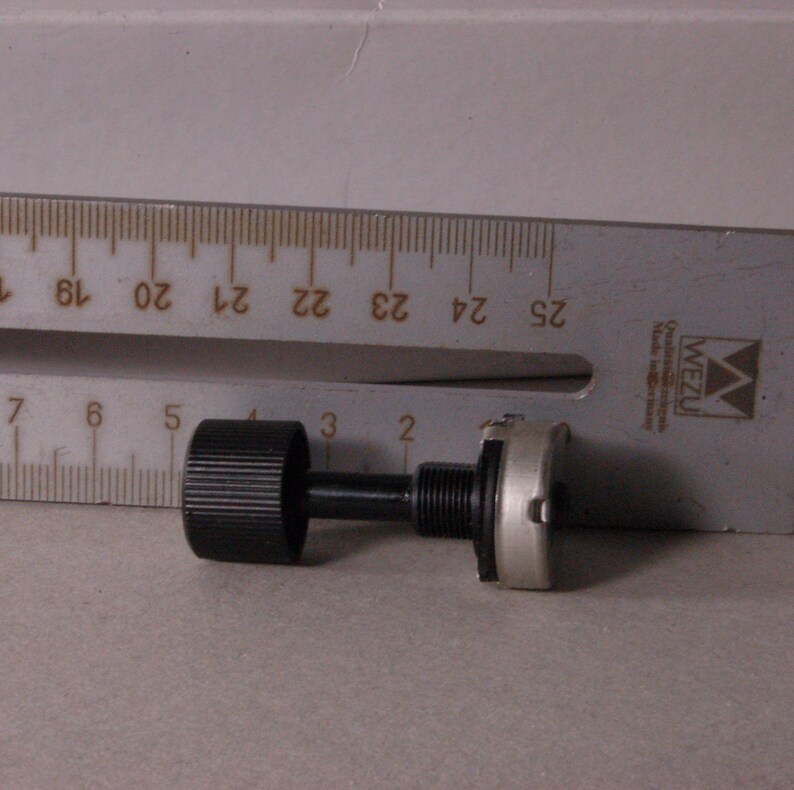Peut inclure: Un bouton de potentiom&egrave;tre noir avec une surface stri&eacute;e, reli&eacute; &agrave; une tige noire et &agrave; une base argent&eacute;e. Une r&egrave;gle m&eacute;tallique avec des marquages en centim&egrave;tres est en arri&egrave;re-plan.