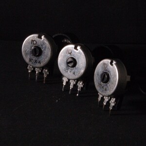 Peut inclure: Trois potentiom&egrave;tres argent&eacute;s avec des boutons noirs et des broches. Chaque potentiom&egrave;tre porte l'inscription "10 K A". Les objets sont dispos&eacute;s en rang&eacute;e sur un fond sombre.