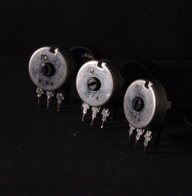 Peut inclure: Trois potentiom&egrave;tres argent&eacute;s avec des boutons noirs et des broches. Chaque potentiom&egrave;tre porte l'inscription "10 K A". Les objets sont dispos&eacute;s en rang&eacute;e sur un fond sombre.