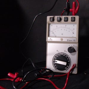 Puede incluir: Un multímetro Elavi 15 vintage con un cuerpo blanco y gris. El dispositivo presenta múltiples diales, medidores y terminales, incluida una escala de voltaje y configuraciones de resistencia. Cables rojos y negros con conectores están adjuntos.