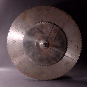 May include: A unique clock made from two circular saw blades. The larger blade forms the clock's face, with a smaller blade nested inside. Black clock hands indicate the time. The metal has a rustic, aged appearance.