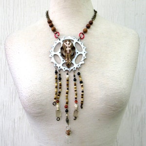 Puede incluir: Un collar de cadena de bicicleta plateado con un colgante de calavera y cuentas de madera marrón. El colgante está rodeado por un engranaje plateado con fondo blanco. El collar tiene múltiples hebras de cuentas en marrón, negro, blanco y rojo.