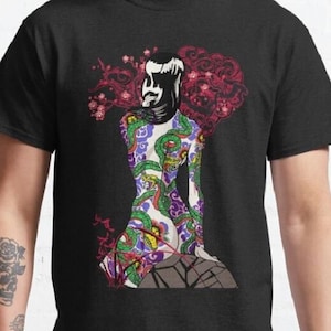May include: A black t-shirt with a colorful illustration of a woman with a snake tattoo. The woman is sitting on a rock with flowers and clouds behind her.