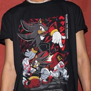 Può includere: Maglietta nera con una grafica di Shadow the Hedgehog della serie di videogiochi Sonic the Hedgehog. La grafica mostra Shadow in una posa di combattimento con uno schema di colori rosso e nero. Il testo "SHADOW THE HEDGEHOG" è stampato sul lato destro della maglietta. Il testo "I'm letting go of that destructive anger. But I will continue to fight, fight, with your wish in my heart. Your wish for everyone to have a chance to be happy" è stampato nella parte inferiore della maglietta.