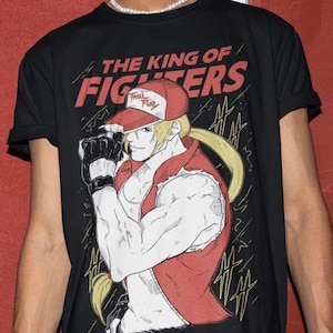 May include: Black t-shirt with a red and white illustration of a character from the video game The King of Fighters. The character is wearing a red vest and a red and white baseball cap with the words "The King of Fighters" printed on the shirt.