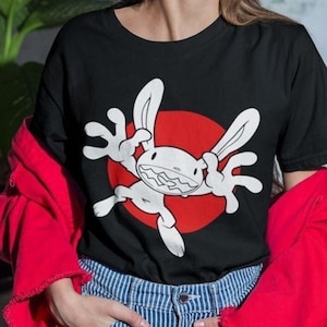 Può includere: Una maglietta nera con un disegno di coniglio bianco a cartone animato che indossa un cerchio rosso intorno a sé. Il coniglio ha un ampio sorriso e alza le braccia in aria.