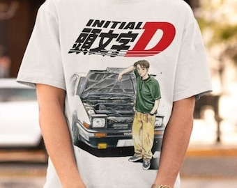 May include: White t-shirt featuring the text "INITIAL D" in black and red, with Japanese characters. The graphic includes an illustration of a car and a person leaning against it. The shirt has short sleeves.