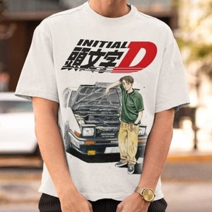 Peut inclure: T-shirt blanc avec l'inscription "INITIAL D" en noir et rouge, ainsi que des caractères japonais. Le graphisme comprend une illustration de voiture et une personne appuyée dessus. Le t-shirt a des manches courtes.
