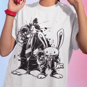 Può includere: Maglietta bianca con una stampa grafica in bianco e nero di due personaggi dei cartoni animati che tengono delle pistole. I personaggi sono un coniglio e un cane che indossa un cappello e un vestito.