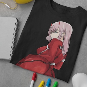 May include: Black t-shirt featuring a graphic print of an anime character with pink hair and a red outfit. The character is holding a bubble in their mouth. The t-shirt is folded and lying on a grey surface with a notebook, headphones, and markers.