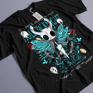 May include: Black t-shirt featuring a detailed graphic of a character with a white head, blue wings, and a sword. The design includes the text "Hollow Knight" in teal, along with other decorative elements. A tag is attached to the shirt.