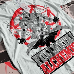 Peut inclure: T-shirt blanc avec un motif graphique de la série animée "Fullmetal Alchemist". Le design présente des personnages en noir et blanc, un symbole circulaire rouge et le titre de la série.