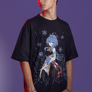 Peut inclure: Un t-shirt noir avec un imprim&eacute; graphique d'un personnage d'anime aux cheveux bleus et une tenue blanche. Le personnage est entour&eacute; de flocons de neige. Le t-shirt est port&eacute; par une personne tenant deux manettes de jeu.