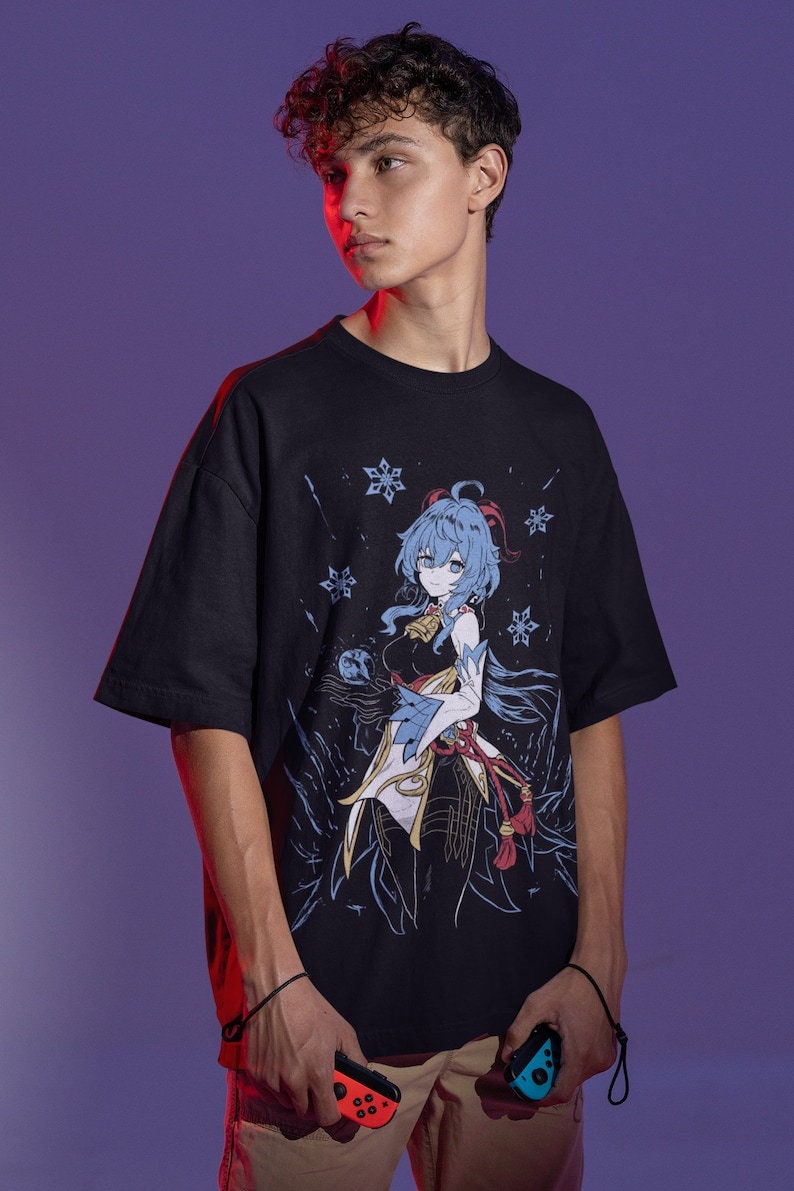 Peut inclure: Un t-shirt noir avec un imprim&eacute; graphique d'un personnage d'anime aux cheveux bleus et une tenue blanche. Le personnage est entour&eacute; de flocons de neige. Le t-shirt est port&eacute; par une personne tenant deux manettes de jeu.