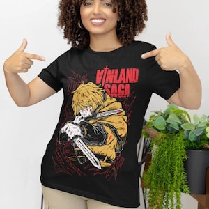 Może przedstawiać: Czarny T-shirt z grafiką postaci z anime "Vinland Saga". Postać trzyma miecz i ma zdecydowany wyraz twarzy. Tekst "Vinland Saga" jest nadrukowany na koszulce na czerwono.