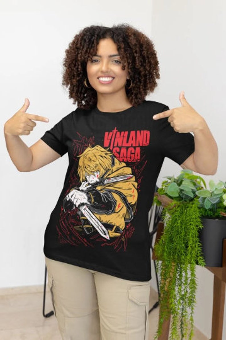 Może przedstawiać: Czarny T-shirt z grafiką postaci z anime "Vinland Saga". Postać trzyma miecz i ma zdecydowany wyraz twarzy. Tekst "Vinland Saga" jest nadrukowany na koszulce na czerwono.