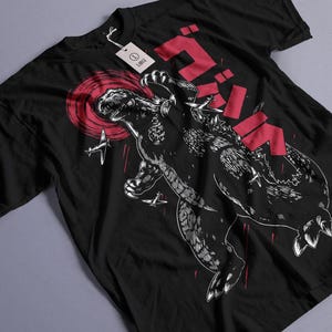 Peut inclure: T-shirt noir avec un motif blanc de Godzilla, un monstre géant, avec le mot japonais "Godzilla" en rouge derrière lui. Le motif comprend également un cercle rouge et des avions.