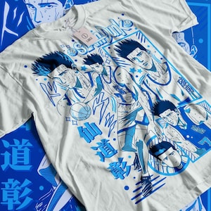 Può includere: Maglietta bianca con una stampa grafica blu e bianca di giocatori di basket. La grafica include il testo "Slam Dunk" e caratteri giapponesi.