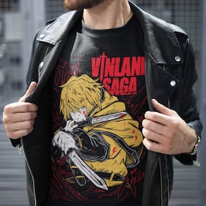 Może przedstawiać: Czarny T-shirt z nadrukiem graficznym blond postaci anime trzymającej miecz. Postać otoczona jest czerwonymi i czarnymi liniami. Tekst "VINLAND SAGA" jest nadrukowany na koszulce czerwonymi literami.
