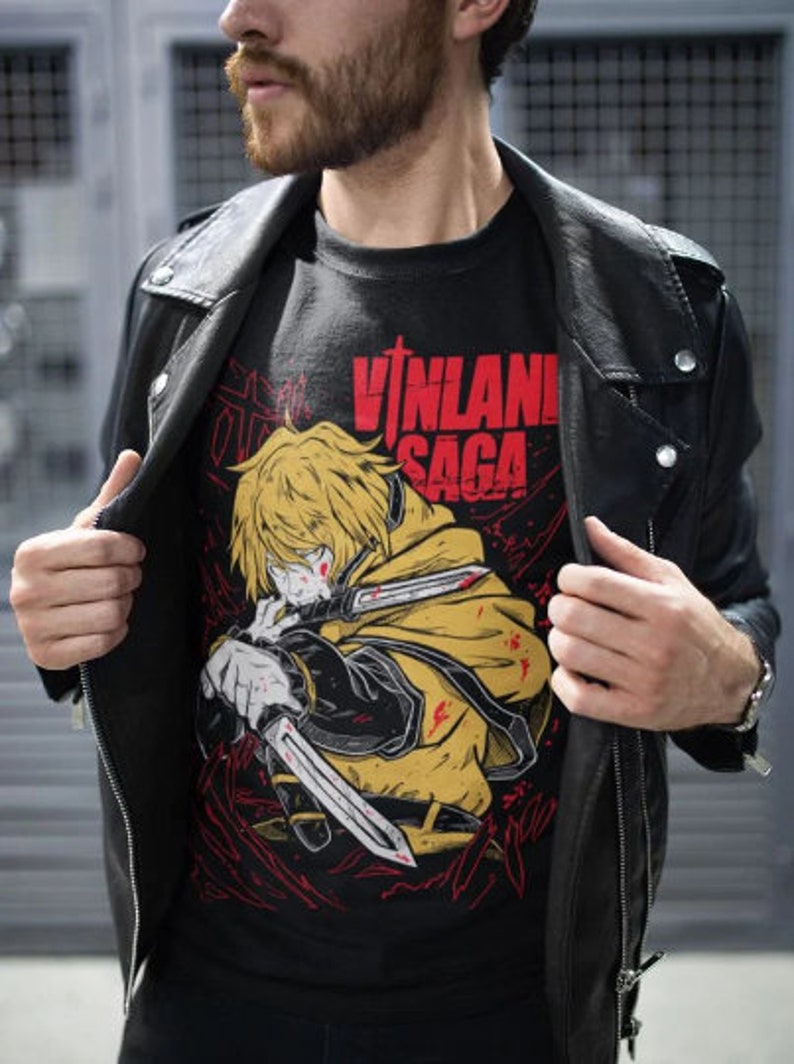 Może przedstawiać: Czarny T-shirt z nadrukiem graficznym blond postaci anime trzymającej miecz. Postać otoczona jest czerwonymi i czarnymi liniami. Tekst "VINLAND SAGA" jest nadrukowany na koszulce czerwonymi literami.