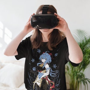 Peut inclure: Un t-shirt noir avec un imprim&eacute; graphique d'un personnage d'anime aux cheveux bleus avec un n&oelig;ud rouge. Le personnage tient un orbe bleu et est entour&eacute; de flocons de neige.