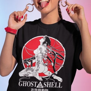 Peut inclure: T-shirt noir avec un graphique circulaire rouge représentant une femme avec des fils et un pistolet. Le texte "Ghost in the Shell" et "攻殻機動隊" est imprimé sous le graphique.