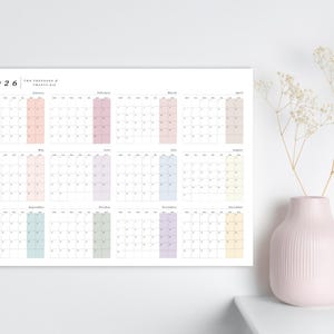 May include: A minimalist 2026 calendar with a white background. Each month is displayed with a pastel color block. A pink vase with white flowers sits to the right.