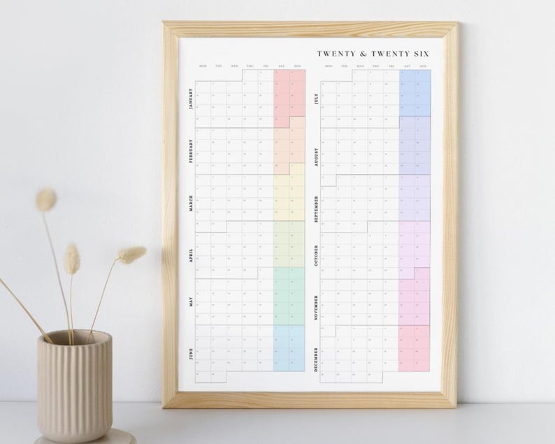 K&ouml;nnte beinhalten: Ein gerahmter Wandkalender mit dem Text "TWENTY & TWENTY SIX" oben. Der Kalender hat ein Rasterlayout mit vertikal aufgelisteten Monaten und horizontalen Daten. Jeder Monat hat einen anderen pastellfarbenen Block.